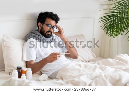 Sick Young Indian Man Looking At Thermometer While Sitting In Bed At Home, Ill Eastern Guy Checking Body Temperature And Touching Aching Head, Suffering Seasonal Flu Symptoms, Copy Space