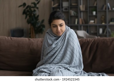 Sick young Indian woman sit on sofa at home wrapped covered in warm duvet or blanket feeling unhealthy ill. Unwell millennial ethnic female feel cold have temperature struggle with flu symptoms.