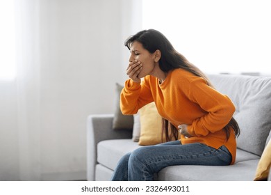 Sick young indian woman sitting on couch, covering her mouth and touching belly, ill lady suffering from food poisoning, home interior, side view, copy space. Foodborne illness
