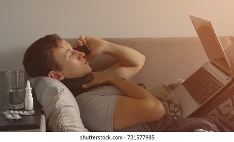 Sick Young Man In Bed Working On Notebook And Talking On The Phone. Plenty Pills And Medications Are On The Bedside Table. Sunlight