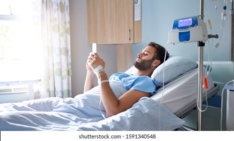 Sick young man lying on a bed in a hospital getting an intravenous drip treatment and using his cellphone - Shutterstock ID 1591340548