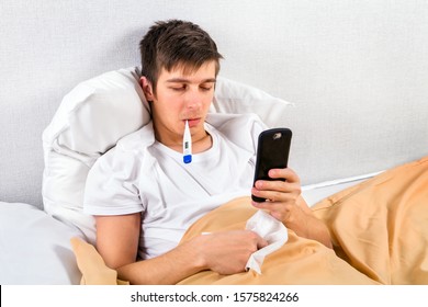 Sick Young Man With A Phone And A Thermometer In The Bed At The Home