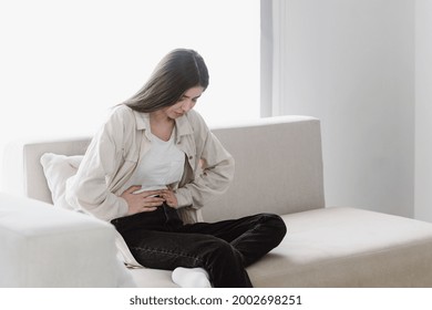 Sick And Young Woman Feeling Stomach Pain, Touch Her Belly, Suffer From Ache, Sitting On Sofa In Living Room Alone. Unhappy Female With Diarrhea Problem And Discomfort In Abdomen