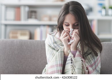 Sick Young Woman At Home On The Sofa With A Cold, She Is Covering With A Blanket And Blowing Her Nose