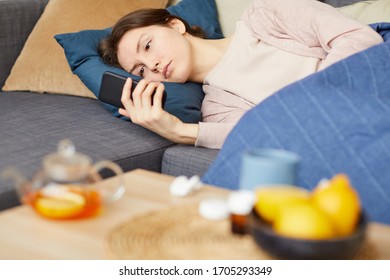 Sick Young Woman Lying On Sofa And Reading A Message On Her Mobile Phone At Home