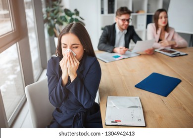 Sick Young Woman Sit Saparately At Table In Meeting Room And Sneezing. She Hold White Napkin. Her Colleagues Work Behind.