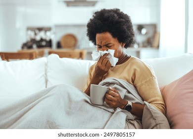 Sick young woman sitting on sofa blowing her nose at home in the sitting room. Photo of sneezing woman in paper tissue. Picture showing woman sneezing on tissue on couch in the living-room - Powered by Shutterstock