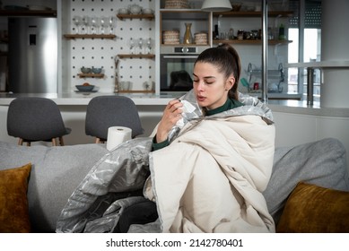 Sick young woman sitting under the blanket on sofa and sneeze with tissue paper at home. Female blowing nose, coughing or sneezing in tissue at home, suffering from flu. Common cold and fever concept.