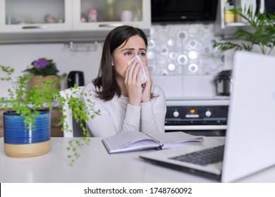 Sick Young Woman. Sneezing And Coughing With Handkerchief, Sitting With Laptop In The Home Office, Kitchen Interior Background. Virus, Seasonal Colds, Allergies, Flu Season