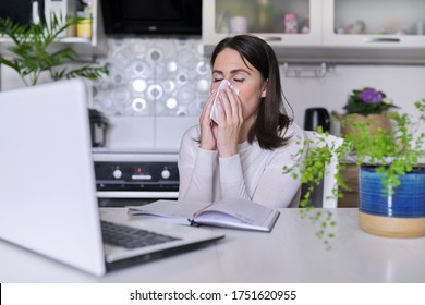 Sick Young Woman. Sneezing And Coughing With Handkerchief, Sitting With Laptop In The Home Office, Kitchen Interior Background. Virus, Seasonal Colds, Allergies, Flu Season