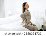 Sick young woman suffering from acute pain in right side, sitting on bed at home, touching lower back with hand, copy space