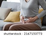 Sick young woman suffering from menstrual pain. Woman with hands squeezing belly having painful stomach ache or period cramps sitting on sofa, Abdominal pain, gastritis, and painful periods concept