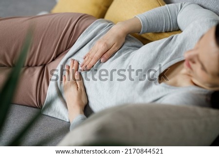 Sick young woman suffering from menstrual pain. Woman with hands squeezing belly having painful stomach ache or period cramps sitting on sofa, Abdominal pain, gastritis, and painful periods concept