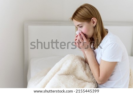 Sick young woman suffers from stuffy nose and sore throat. Upset sick Caucasian lady sitting in bed blowing her nose using a paper napkin. Cold and flu concept. A sick young woman coughs, sneezes Сток-фото © 