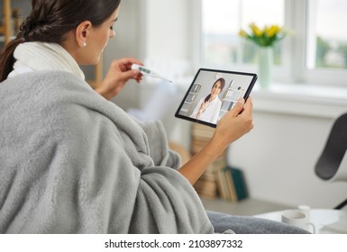 Sick young woman who has high temperature is using digital tablet device, having online telemedicine consultation, holding thermometer, telling doctor about symptoms, asking for help and prescription