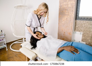 Side angle full length portrait of pretty female cosmetologist making mask procedures for her charming African girl patient, using brush and treatment gel mask.