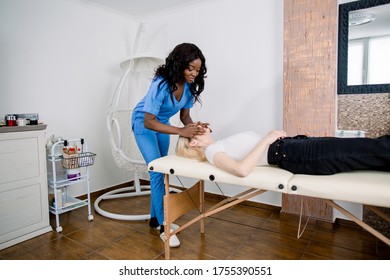 Side angle full length portrait of pretty African woman cosmetologist making face massage for her charming Caucasian girl patient, in modern beauty center