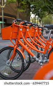 Side Back to Front Vertical View, Line Up of vibrant Orange Rental System Sharing Station Loaner Bikes Bicycles Tires, Seats, Baskets, City Background Shallow Field of Depth - Portland, Oregon