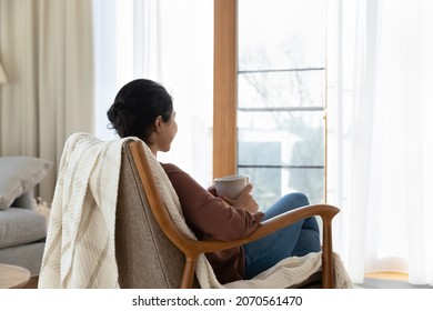 Side back view calm Indian woman relax on comfy lounge armchair with tea cup enjoy morning at modern apartment. Peaceful female daydreaming looks out window, stress-free, favourite beverage concept
