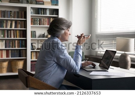 Side back view concentrated middle aged old businesswoman recording audio message on smartphone, communicating with partners sharing ideas in loudspeaker conversation, discussing online project.