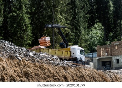 Side Bottom Pov Big Modern Truck With Mounted Self Loading Crane Arm Boom Unloading Pallet Of New Bricks At Countryside Rural Forest Suburban Construction Site. Building Materials Delivery Service