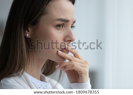 Side close up head shot view nervous anxious young woman standing near window, thinking of personal or health problems, feeling doubtful about hard decision, suffering from loneliness indoors.