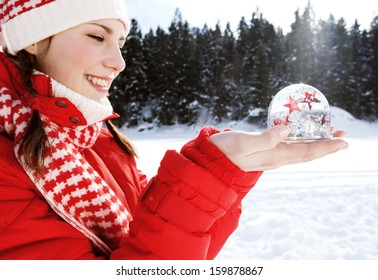 Side close up portrait of a joyful young woman holding a snow globe with red and white glitter stars floating inside, while in the snow mountains during a winter vacation, outdoors. - Powered by Shutterstock