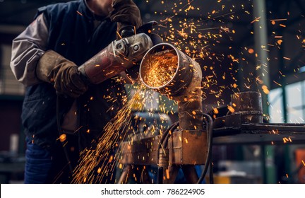 Side close up view of professional focused worker man in uniform working on the metal pipe sculpture with an electric grinder while sparks flying in the industrial fabric workshop. - Powered by Shutterstock