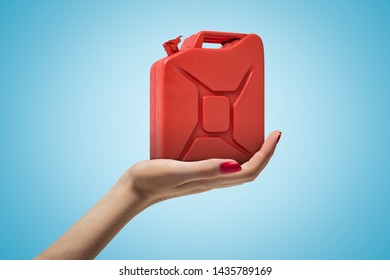 Side closeup of woman's hand facing up and holding red metal jerrican on light blue gradient background. Petroleum industry. Chemical products. Fuel containers.
