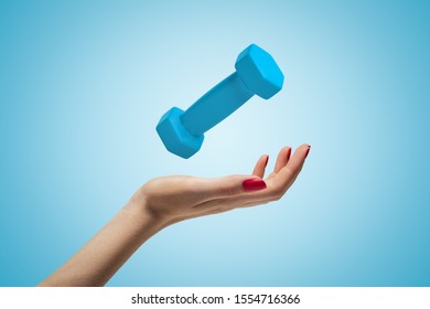 Side Closeup Of Woman's Hand Facing Up And Levitating Small Bright-blue Dumbbell On Light-blue Gradient Background. Pick Up The Right Weight. Work Out. Keep Fit.