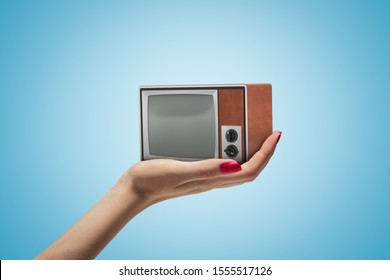 Side Closeup Of Woman's Hand Facing Up And Holding Small Retro TV Set On Light Blue Gradient Background. Retro Technology. Analogue Television. Nostalgic Mood.