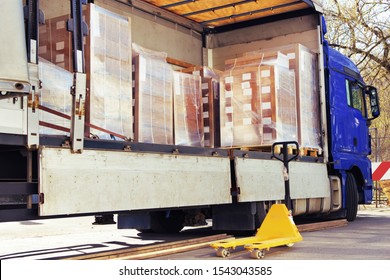 The Side Curtain Of The Trailer Is Opened And The Truck Is Full Of Pallets With Merchandise. There Are Cardboard Boxes Wrapped With Stretch Wrap, The Pallet Jack Is Waiting Beside To Unload The Cargo.