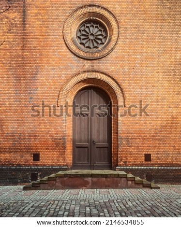 Side facade entrance into catholic church. Veranda porch with a wooden door and round rosette window above it.