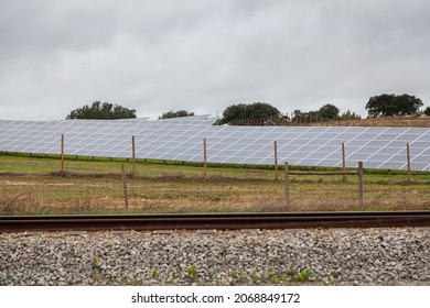 Side front view of multiple solar panels set at angle in green and brown autumn field behind railroad with wire fences on sides under grey sky in rainy day in Portugal