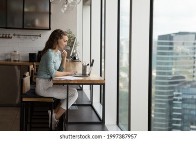 Side Full Length View Happy Concentrated Young Business Lady Working On Computer, Sitting At Table In Modern Home Office Kitchen Room With Panoramic Cityscape View Window, Casual Workday Concept.