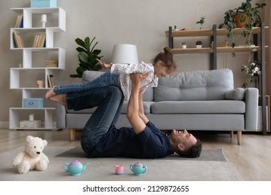 Side Full Length View Laughing Candid Young Father Carrying Little Cute Child Daughter, Lying On Floor Carpet. Happy Two Generations Family Having Fun, Enjoying Entertaining Activity Together At Home.