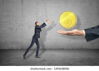 Side Full Length View Of Little Businessman Reaching Out For Big Gold Coin Levitated Above Huge Hand Emerging From Right. Greed For Gain. Earning Money. Gaining Profits.