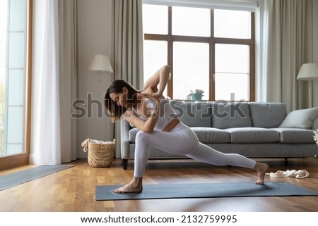Side full-length view young slim woman in white comfy activewear standing barefoot on yogamat perform Crescent Lunge on Knee Prayer Hands asana or Parivrtta Anjaneyasana. Yoga practice at home concept