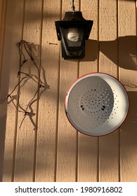 Side Of House With Porch Light, Bent Piece Of Metal Fencing, And Old White And Red Colander Hanging On The Wall In Early Morning Light