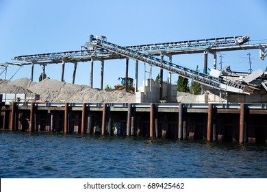 Side Industrial Canal River Scene Of Concrete Manufacturing Facility With Overhead Mechanical Conveyor Belt System Dumping Large Piles Of Sand And Gravel Beside Cement Wall And Rusted Pylon Sea Wall