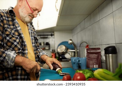 Side low angle view of senior happy man chopping and cutting organic vegetables with knife on counter in kitchen to cook delicious healthy dinner. Grandpa preparing breakfast for his family