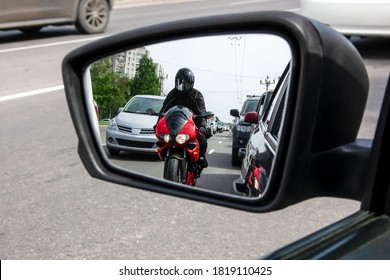The side mirror of the car reflects a motorcyclist who is moving between cars very close to them