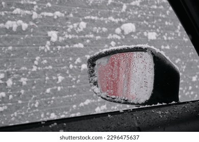A side mirror is seen covered in wet snow through an open car window during a snowfall.