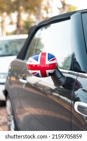 Side Mirror With The United Kingdom Flag. Car Parked In The Street With The UK Flag On The Rear View Mirror. Reflections Of Light On The Bodywork And Windows Of A Car Parked In The Street.