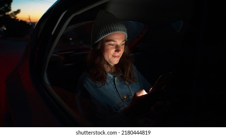 Side night cinematic view of young beautiful woman sit in taxi car back seat near window on road trip, hold, check on mobile phone, use smartphone, scroll social media news, read messages and smile
