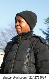 Side On View Of Black Woman In A Black Hat And Black Duvet Coat Looking Off To The Left With Trees In Soft Focus Against A Cold Blue Sky