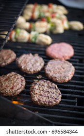 Side Overhead View Of Homemade Beef Hamburger Patties Cooking Over Orange Gas Flame Of Barbecue Smoker With Gourmet Charred Grill Marks And Blurry Chicken Kabab Background At Backyard Summer Cookout