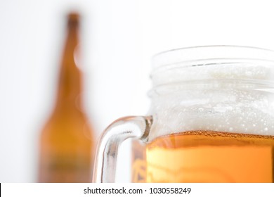 Side perspective close up on a mug of craft ale in a mason jar with a glass handle, and a brown beer bottle in bright white background