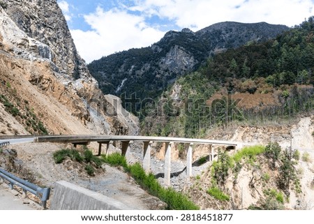 Side perspective of a concrete bridge built along a steep, rocky mountain cliff face with dense forest slopes under a vibrant sky in Yunnan, China.