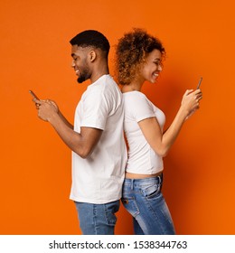 Side Portrait Of African Couple Standing Back To Back With Mobile Phones, Orange Studio Background